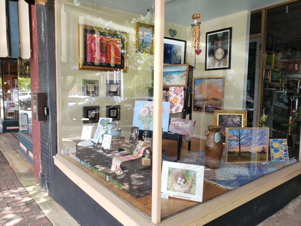 an outside view of the Madison Artist Guild and MAGallery window showing paintings, wind chimes, pottery, and other artwork for sale